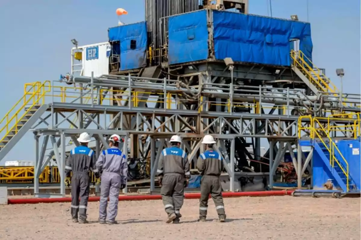 Four workers wearing YPF helmets and uniforms observe an industrial structure at an oil site. Four workers wearing YPF helmets and uniforms observe an industrial structure at an oil site.