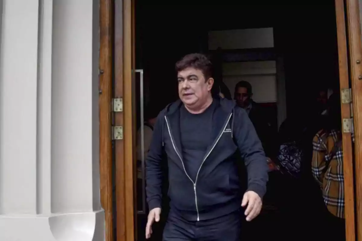 Hombre de cabello oscuro con chaqueta negra saliendo por una puerta de madera mientras otras personas permanecen en el interior Hombre de cabello oscuro con chaqueta negra saliendo por una puerta de madera mientras otras personas permanecen en el interior