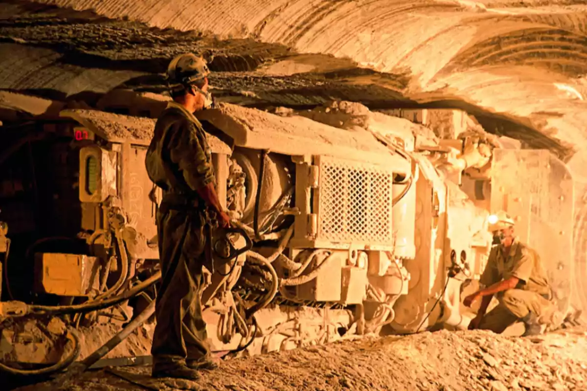 Two workers wearing helmets and safety gear operate heavy machinery inside an underground mine illuminated by artificial light. Two workers wearing helmets and safety gear operate heavy machinery inside an underground mine illuminated by artificial light.