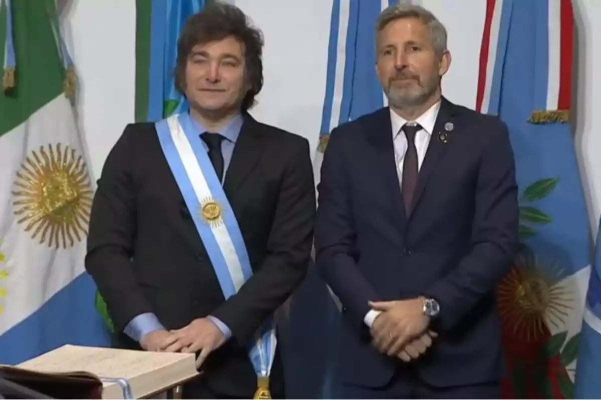 Dos hombres de traje posan frente a banderas argentinas uno de ellos lleva una banda presidencial y una medalla dorada