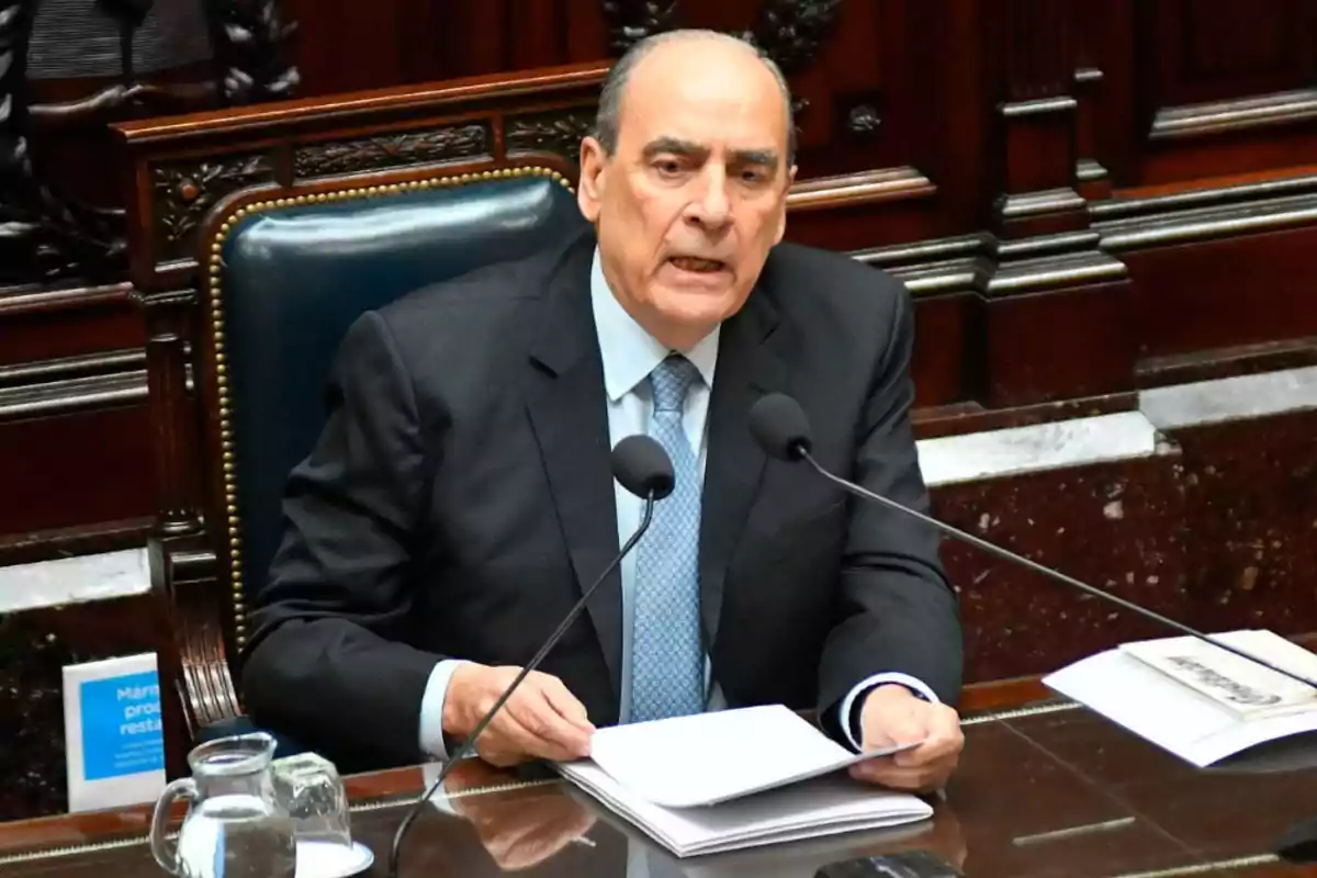 Man in a suit speaking in front of two microphones in a government room with documents on the table Man in a suit speaking in front of two microphones in a government room with documents on the table