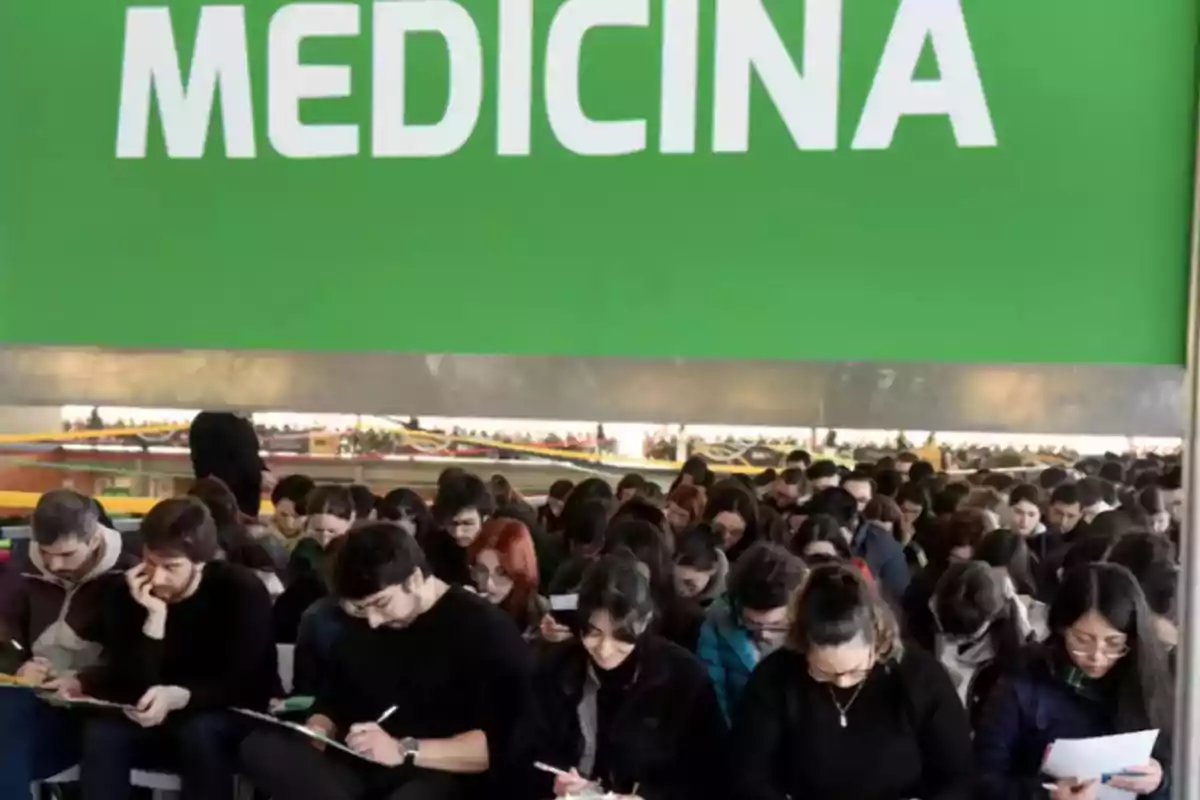 Un gran grupo de estudiantes concentrados escribiendo bajo un letrero verde que dice MEDICINA