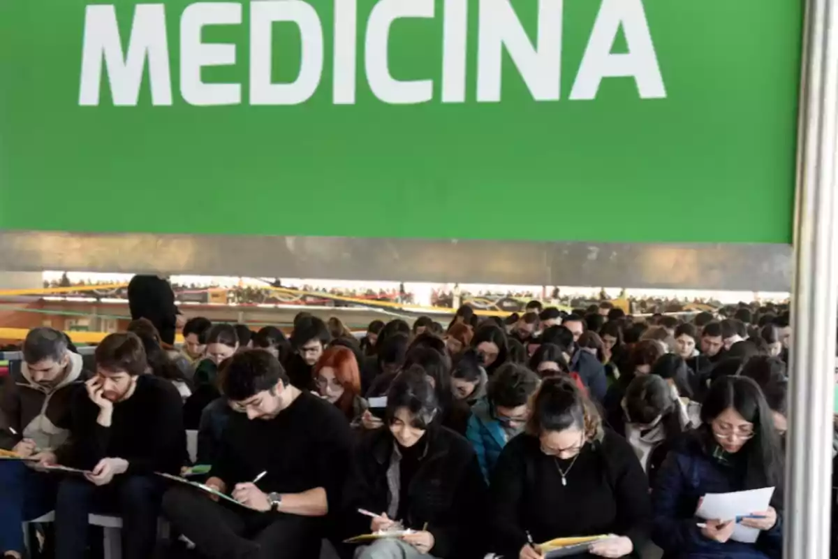 Un gran grupo de estudiantes concentrados resolviendo exámenes bajo un cartel verde que dice MEDICINA