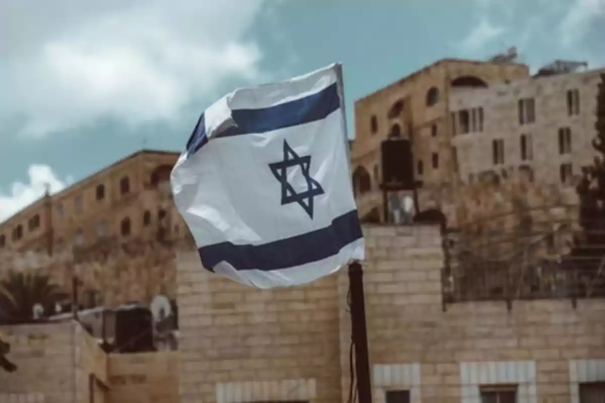 Israeli flag waving in front of stone buildings under a partly cloudy sky