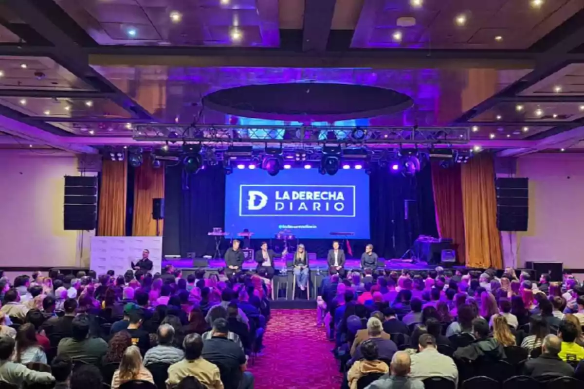 Auditorium full of people attending an event with a panel of six people on stage and a large screen in the background displaying the La Derecha Diario logo.