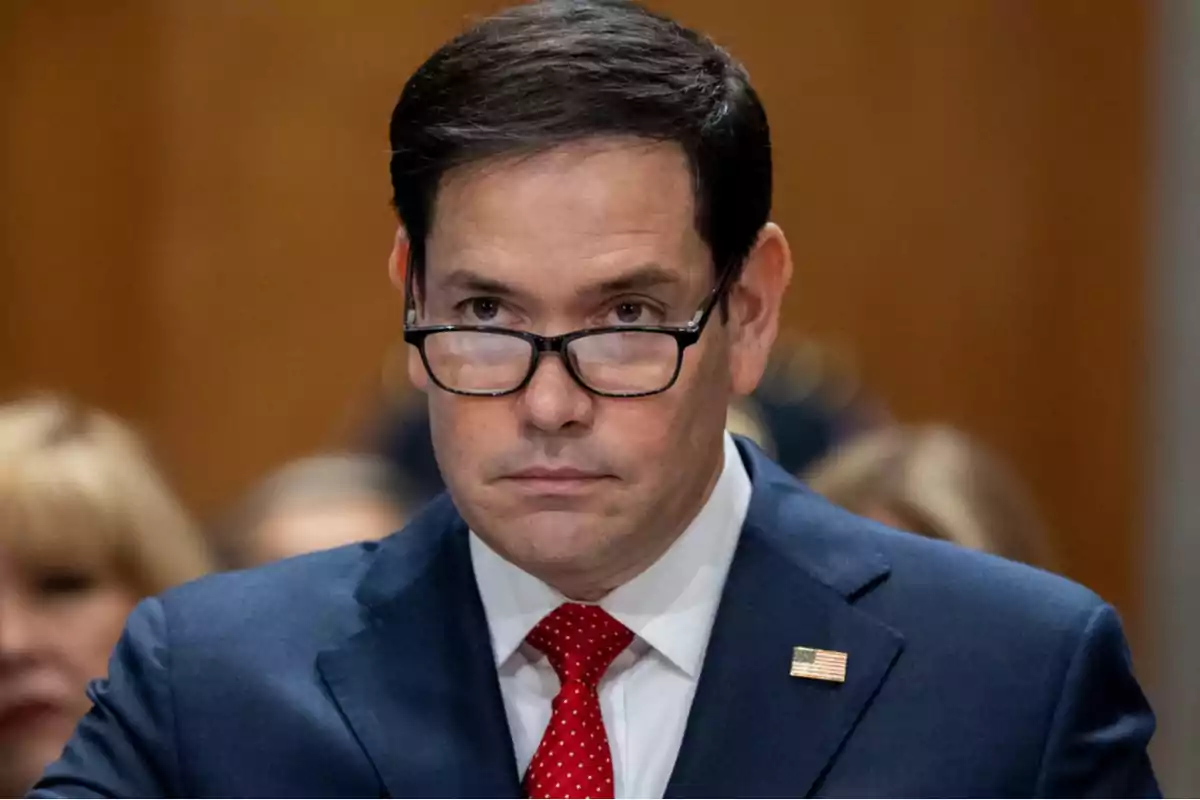 Dark-haired man with glasses and a blue suit, wearing a red tie with white dots and a U.S. flag pin, in a formal setting