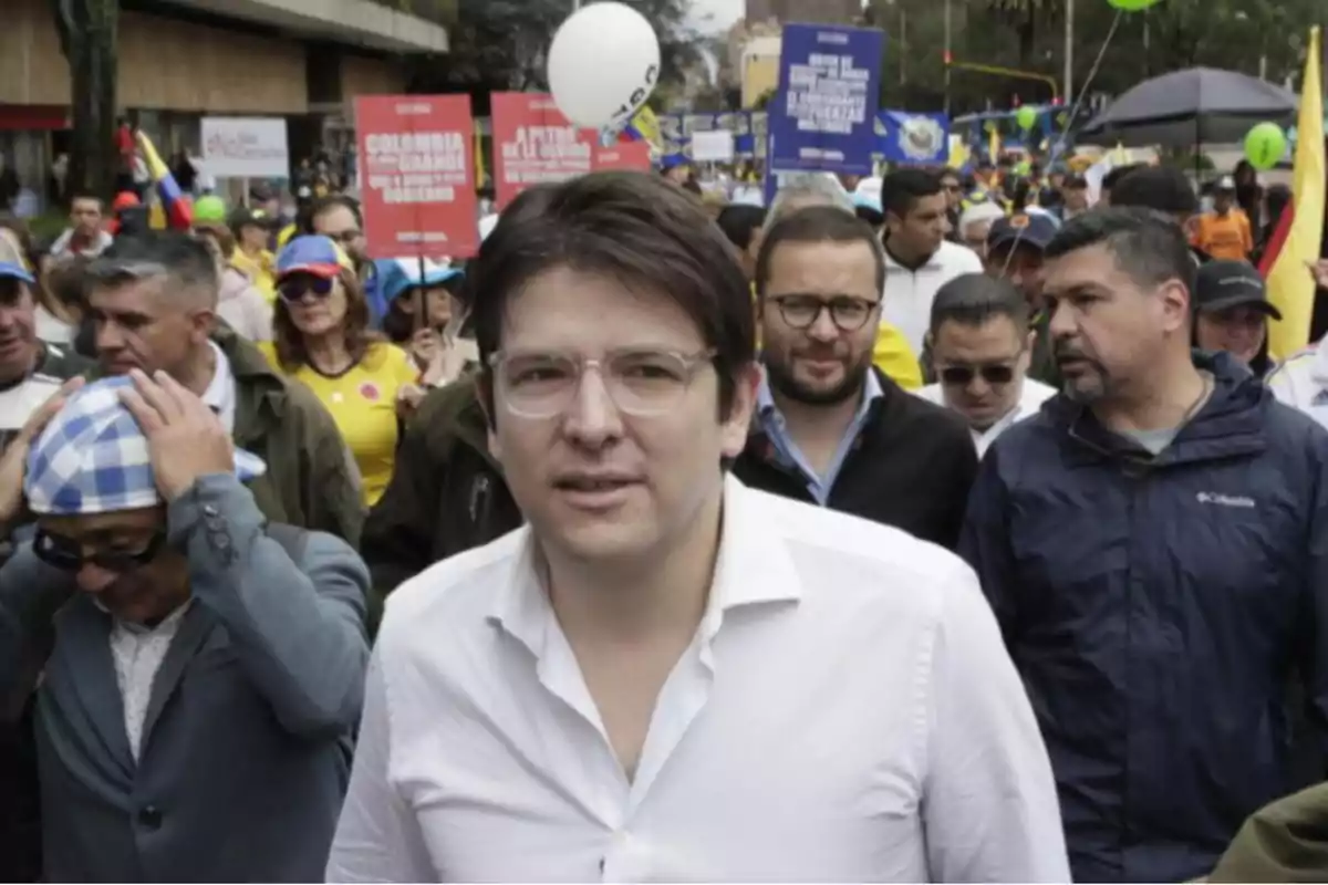 Hombre con gafas y camisa blanca caminando al frente de una multitud durante una manifestación con pancartas y globos