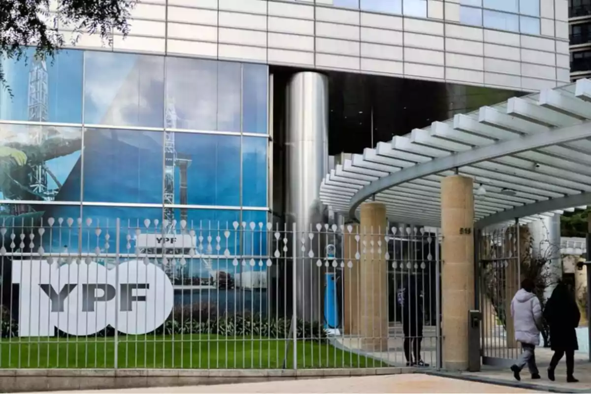 Modern YPF building with a sign celebrating the company’s 100th anniversary and two people walking near the main entrance Modern YPF building with a sign celebrating the company’s 100th anniversary and two people walking near the main entrance
