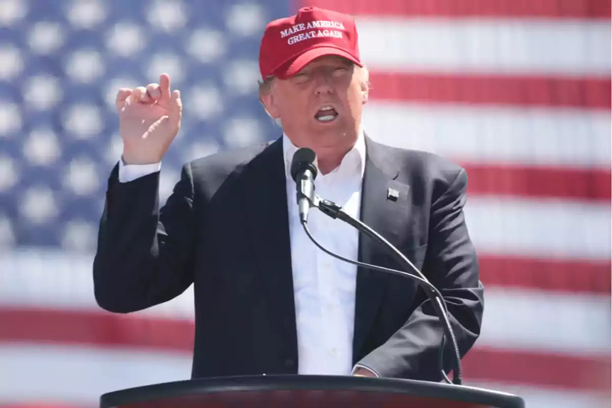 Hombre con gorra roja hablando en un podio frente a una bandera de Estados Unidos