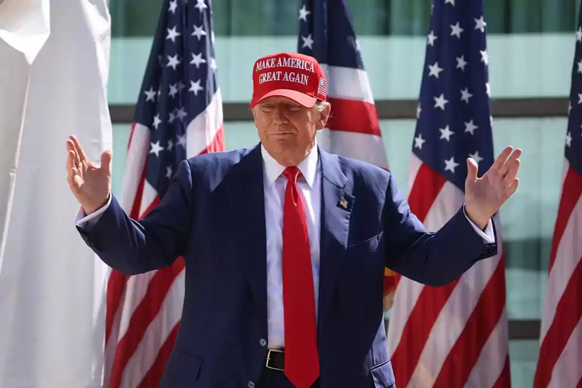 Hombre con traje azul y corbata roja usando una gorra roja con texto blanco, de pie frente a varias banderas de Estados Unidos con los brazos extendidos