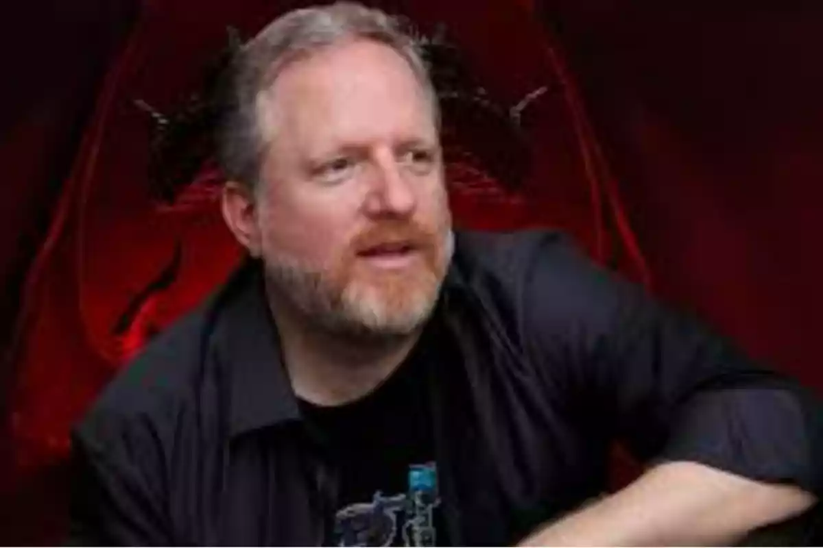 Hombre de barba y cabello claro con camisa oscura sentado frente a un fondo rojo oscuro