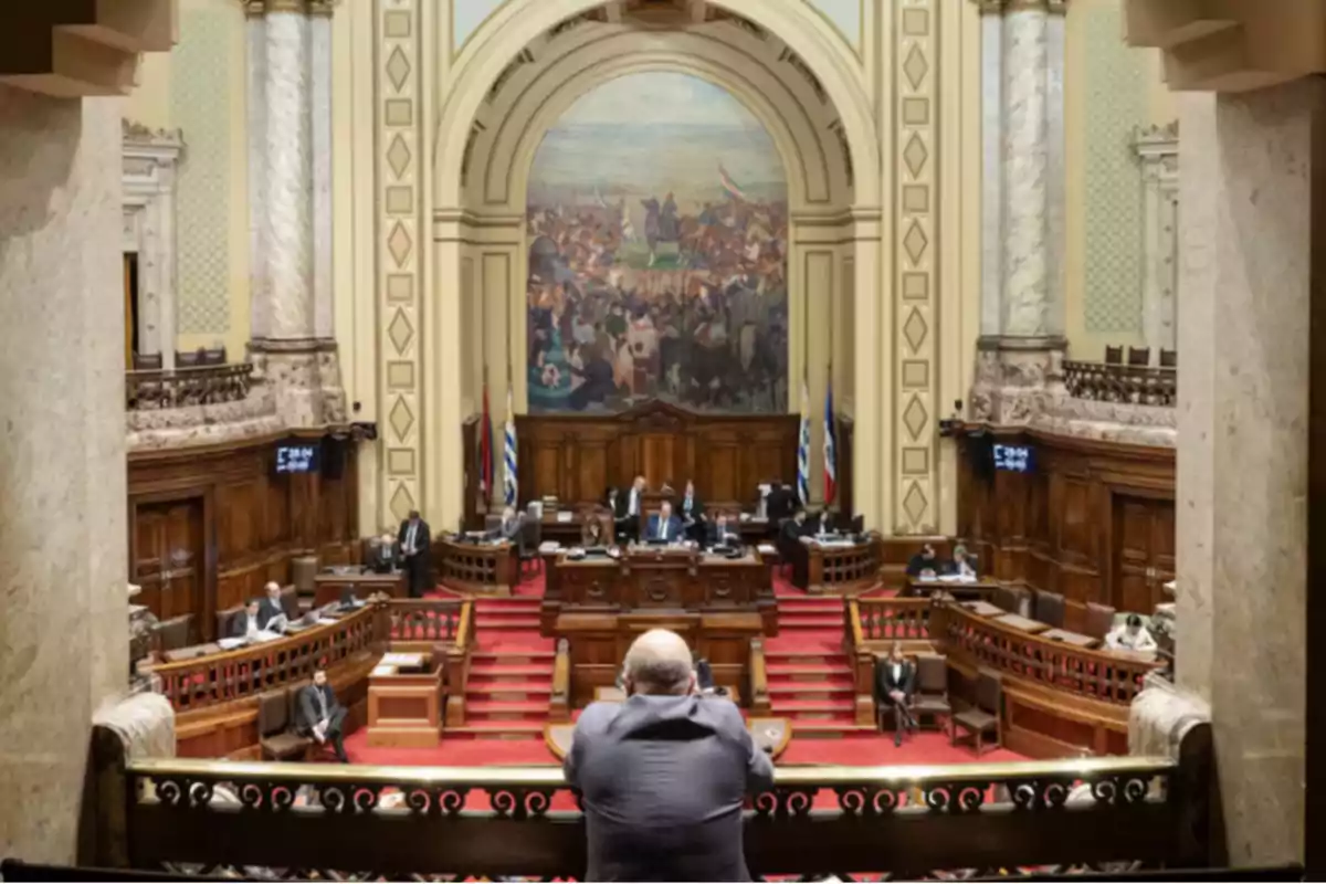 Vista interior de un parlamento con varias personas sentadas en sus bancas y un gran mural al fondo Vista interior de un parlamento con varias personas sentadas en sus bancas y un gran mural al fondo