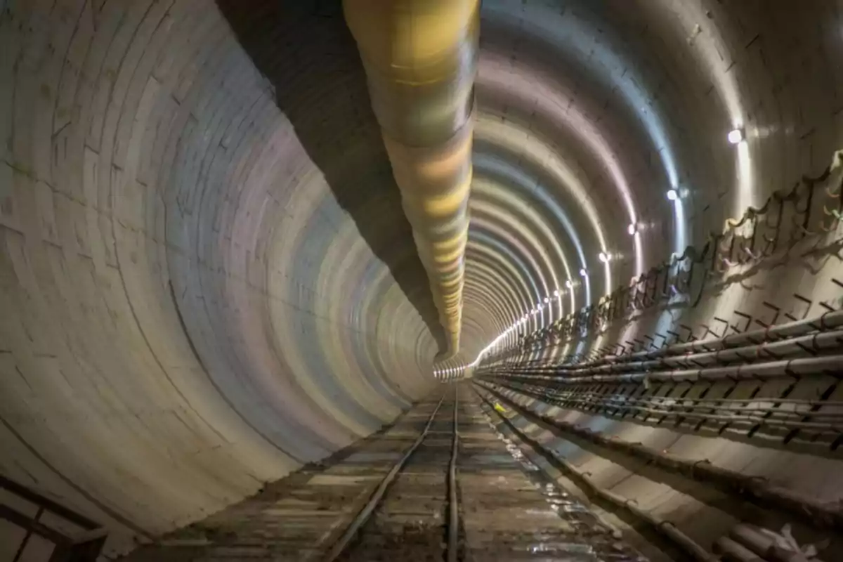 Interior of an underground tunnel with rails and pipes on the sides, illuminated by lights on the wall Interior of an underground tunnel with rails and pipes on the sides, illuminated by lights on the wall