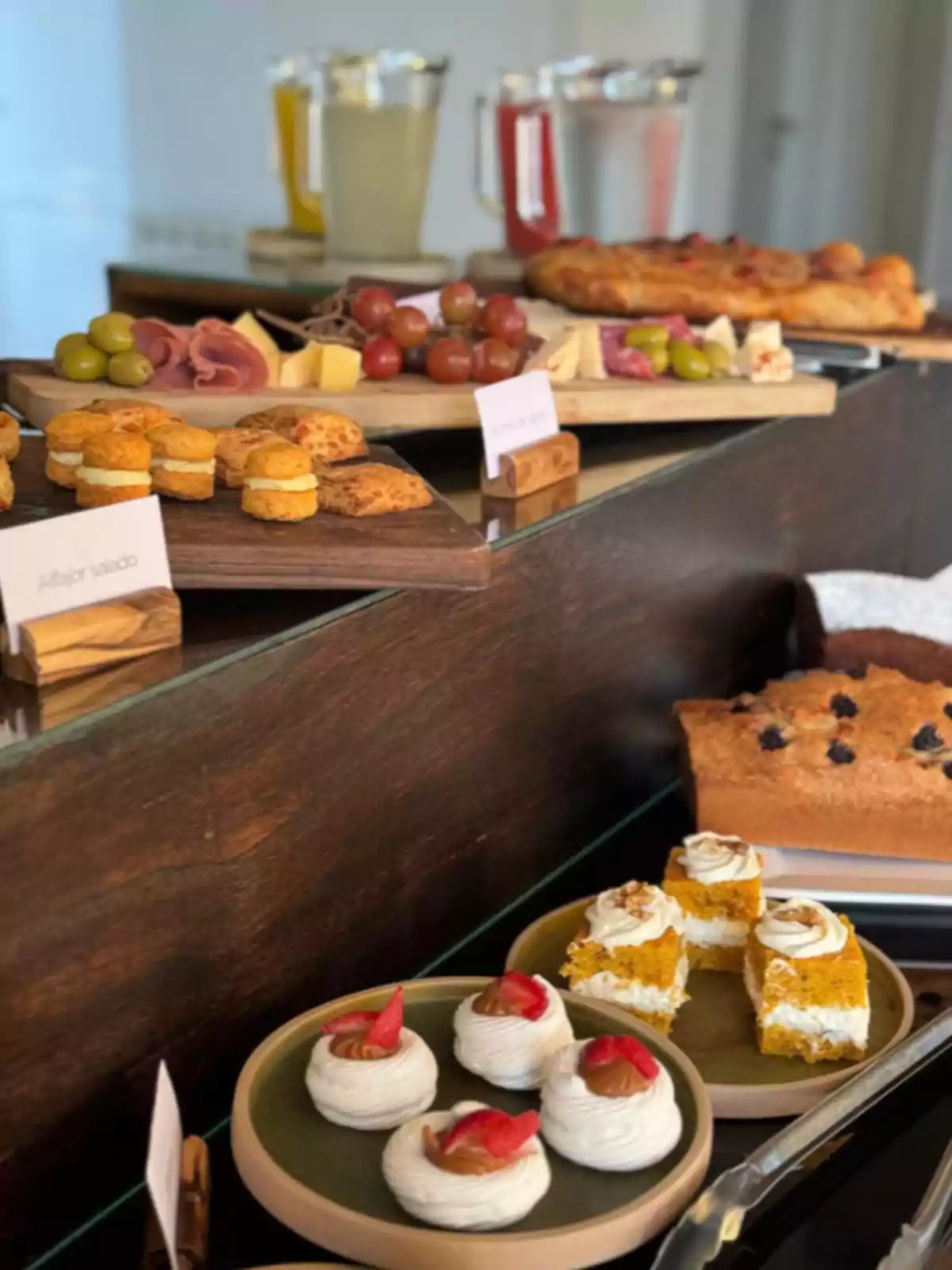 Mesa con variedad de bocadillos dulces y salados, incluyendo alfajores, merengues con dulce de leche, pastel de zanahoria, tabla de quesos, uvas, aceitunas, fiambres y jarras de bebidas al fondo Mesa con variedad de bocadillos dulces y salados, incluyendo alfajores, merengues con dulce de leche, pastel de zanahoria, tabla de quesos, uvas, aceitunas, fiambres y jarras de bebidas al fondo