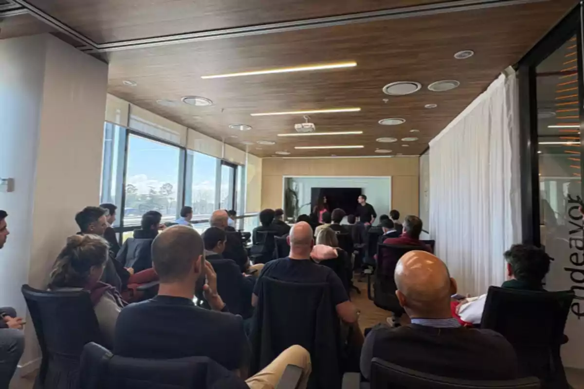 People sitting in a conference room listening to two presenters at the front, with large windows letting in natural light. People sitting in a conference room listening to two presenters at the front, with large windows letting in natural light.