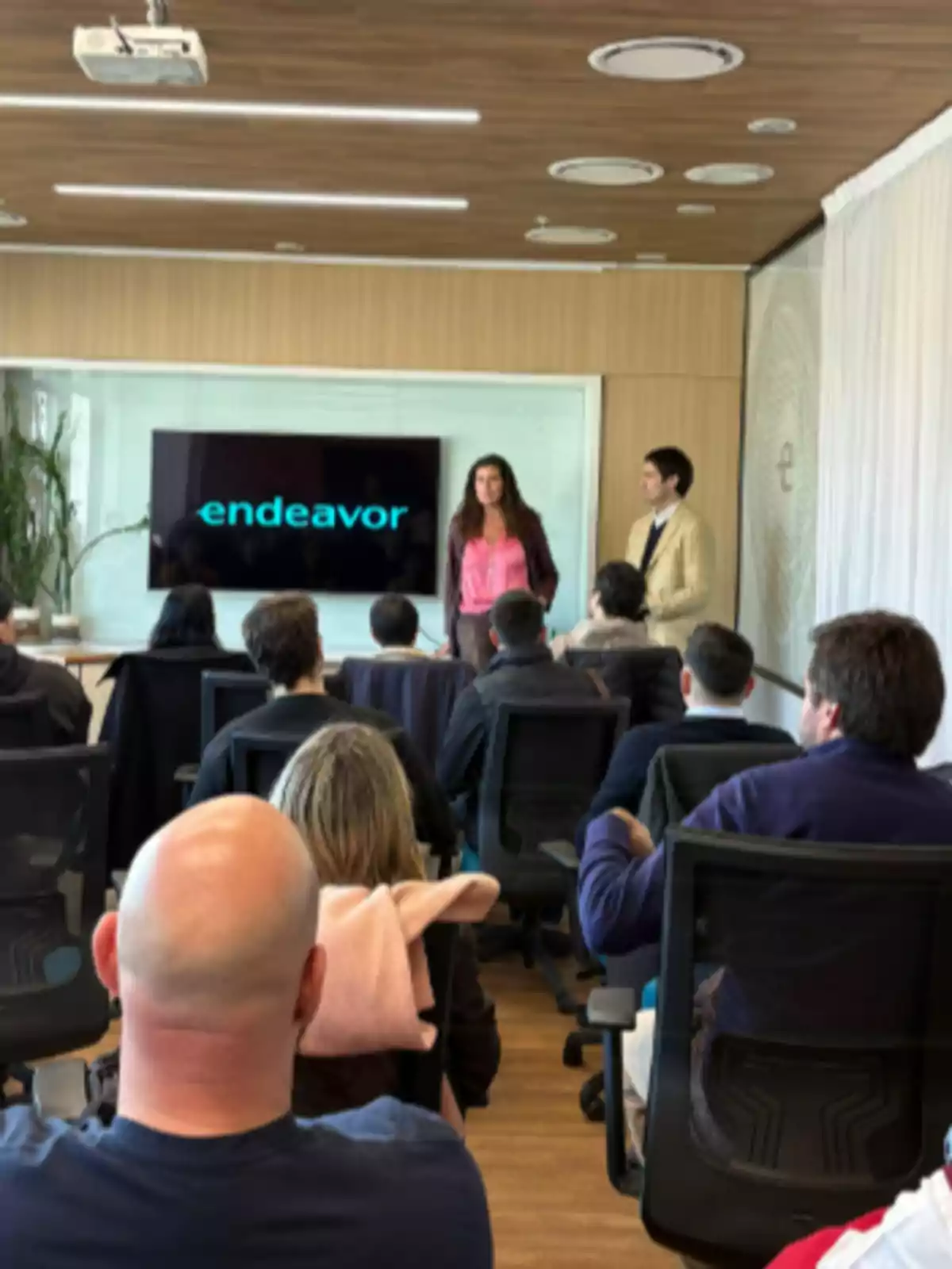 People seated in a conference room while two presenters speak in front of a screen displaying the word People seated in a conference room while two presenters speak in front of a screen displaying the word
