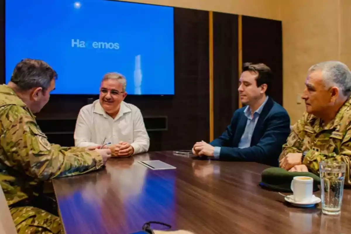Cuatro hombres, dos con uniforme militar y dos con ropa civil, conversan sentados alrededor de una mesa en una sala de reuniones con una pantalla azul encendida al fondo.
