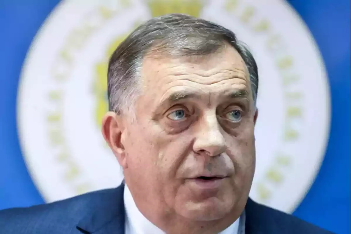 Hombre de mediana edad con cabello corto y canoso usando traje oscuro frente a un fondo azul con un emblema circular desenfocado Hombre de mediana edad con cabello corto y canoso usando traje oscuro frente a un fondo azul con un emblema circular desenfocado