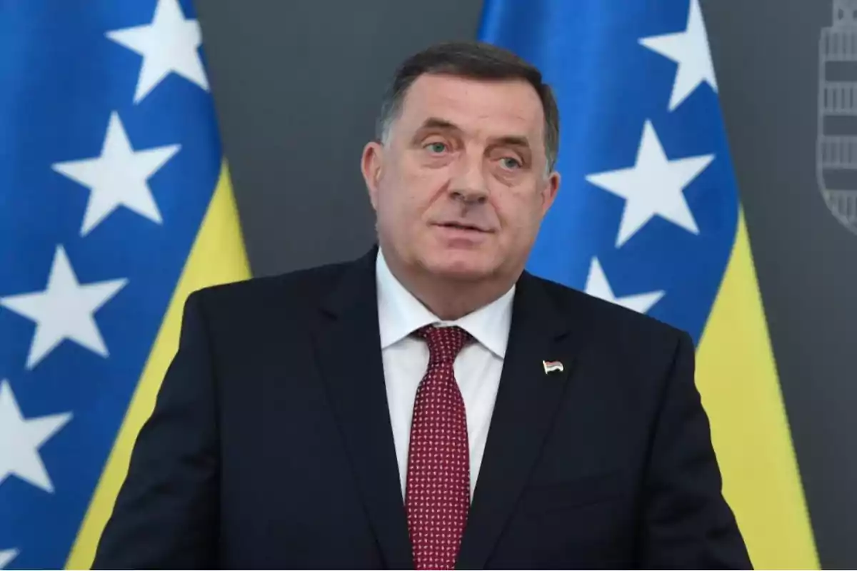 Hombre de traje oscuro y corbata roja posando frente a dos banderas de Bosnia y Herzegovina