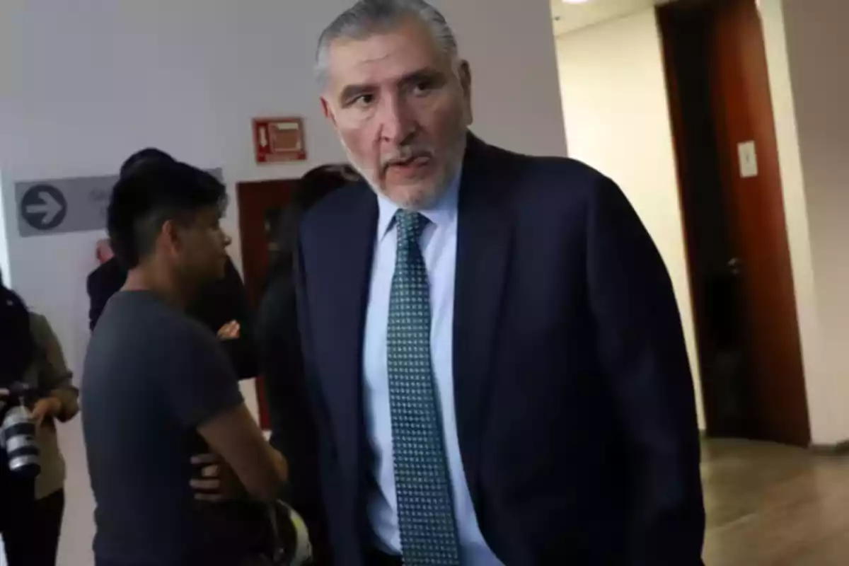 Hombre de cabello canoso y barba con traje oscuro y corbata verde en un pasillo junto a otras personas