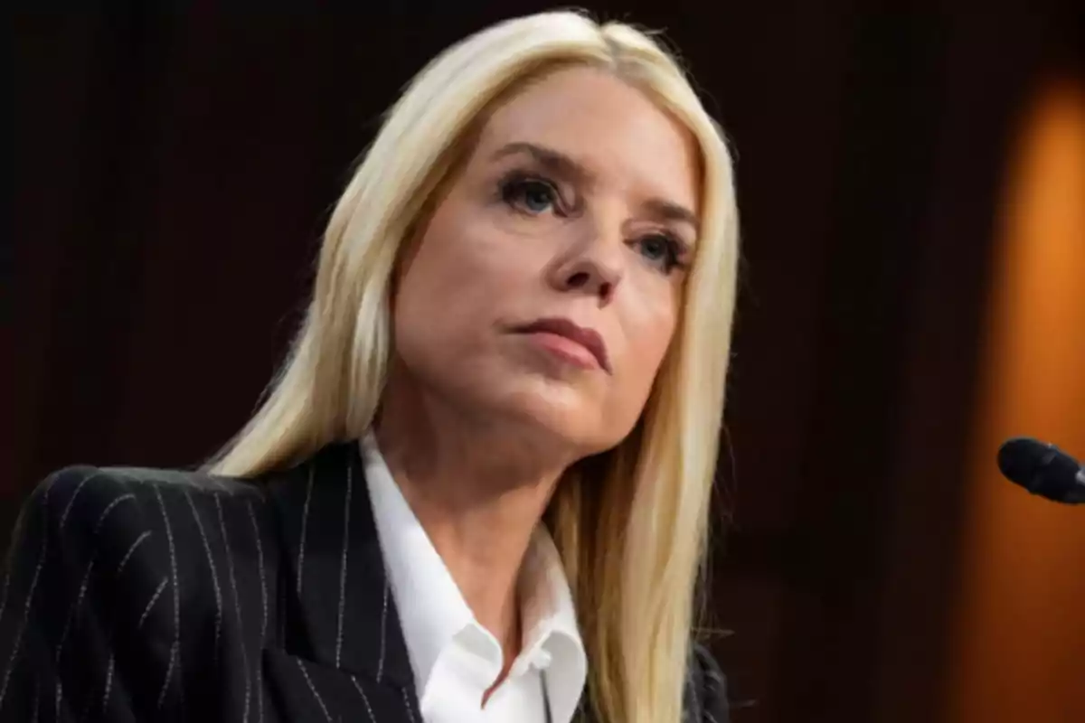 Mujer rubia con expresión seria usando saco oscuro y camisa blanca frente a un micrófono