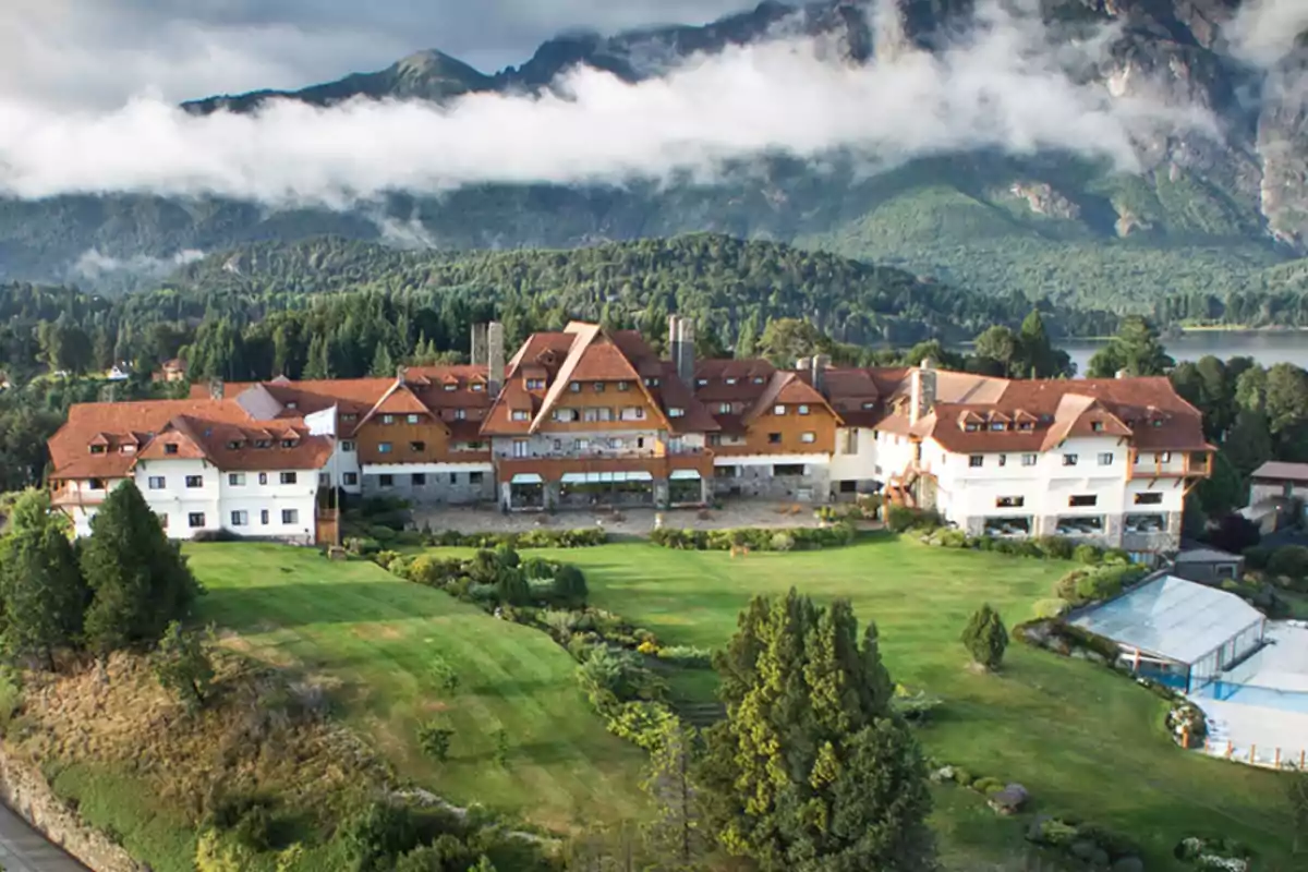 Hotel de estilo alpino rodeado de jardines verdes y montañas boscosas con nubes bajas en el fondo