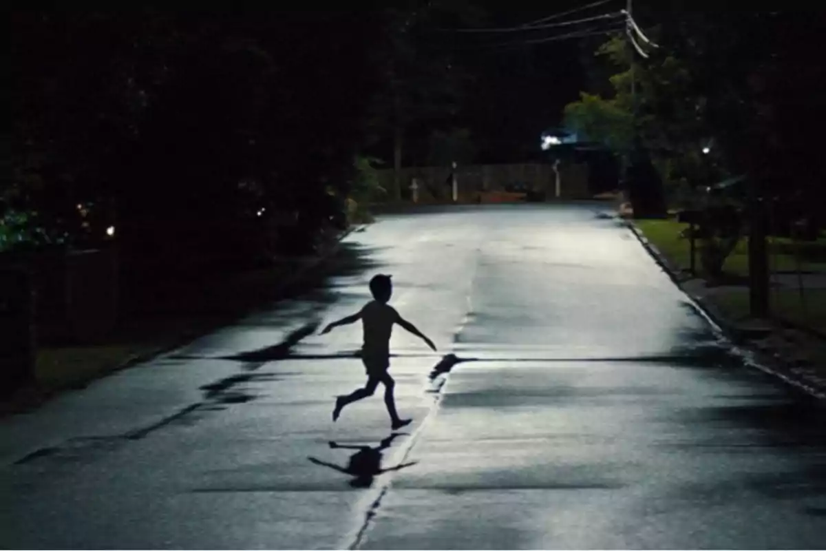 Un niño corre descalzo por una calle vacía y mojada durante la noche iluminado por la luz de una farola