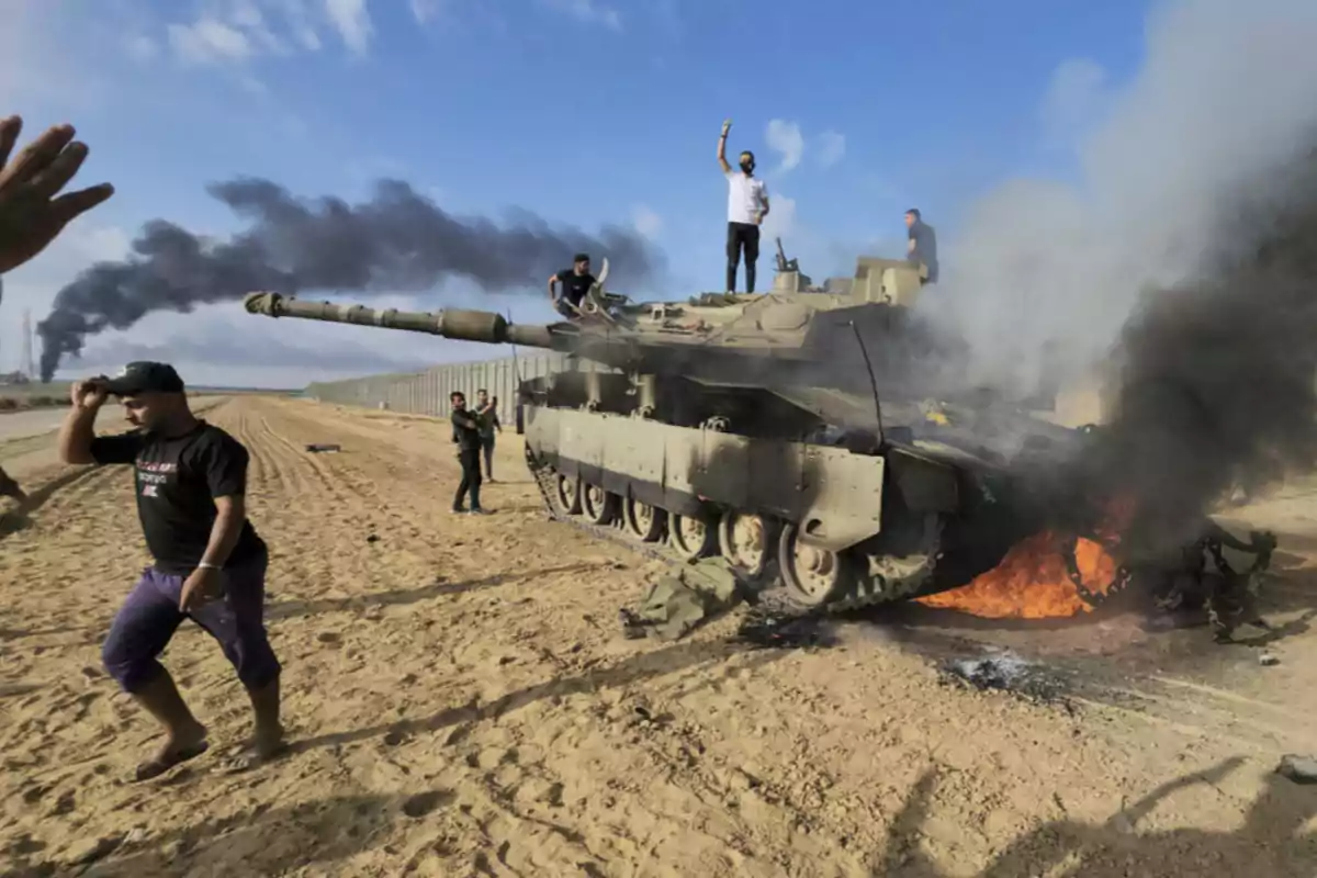 Several people surround and climb onto a burning tank on sandy ground as a column of black smoke rises in the background under a blue sky.