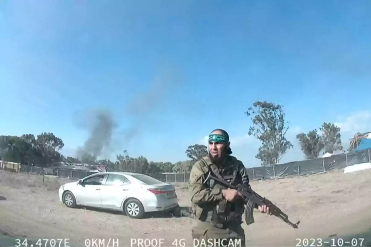 Armed man in military uniform with a green band on his head walking in front of a white car in a rural area with black smoke in the background Armed man in military uniform with a green band on his head walking in front of a white car in a rural area with black smoke in the background