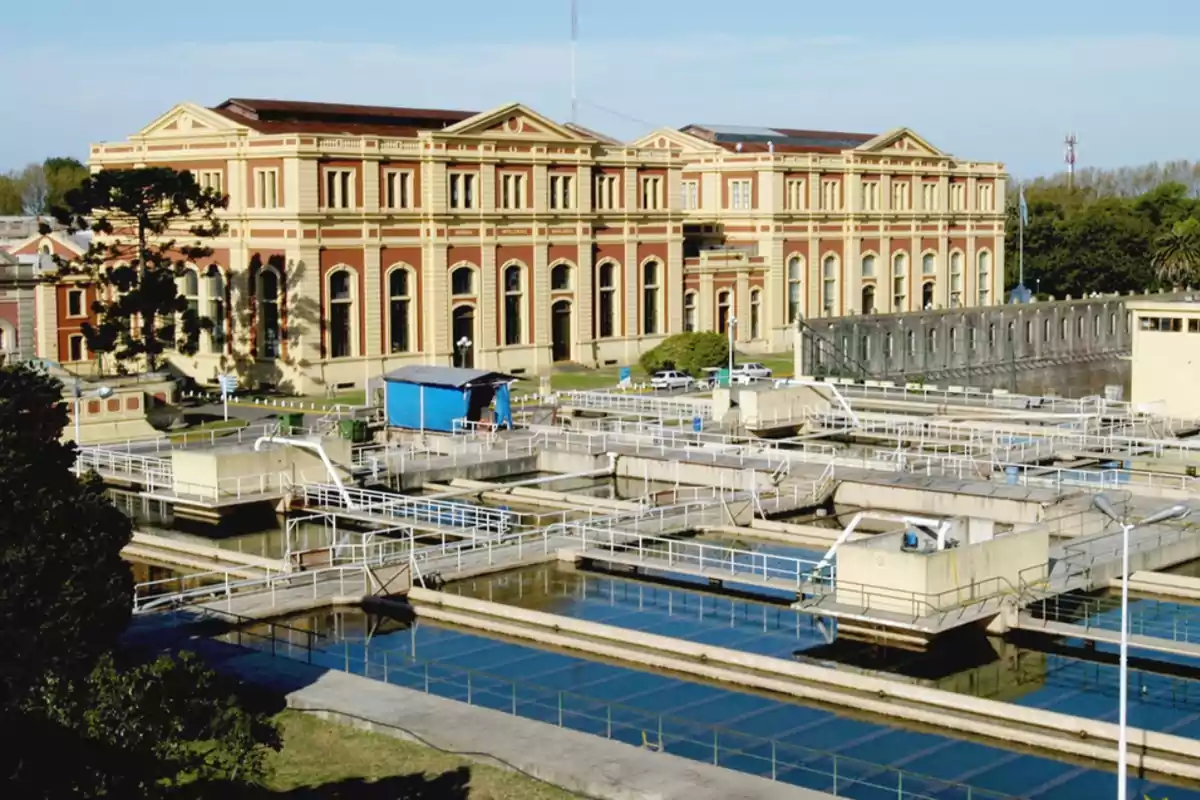 Edificio antiguo de gran tamaño con fachada de ladrillo y detalles en color claro, rodeado de instalaciones modernas de tratamiento de agua con tanques y pasarelas blancas