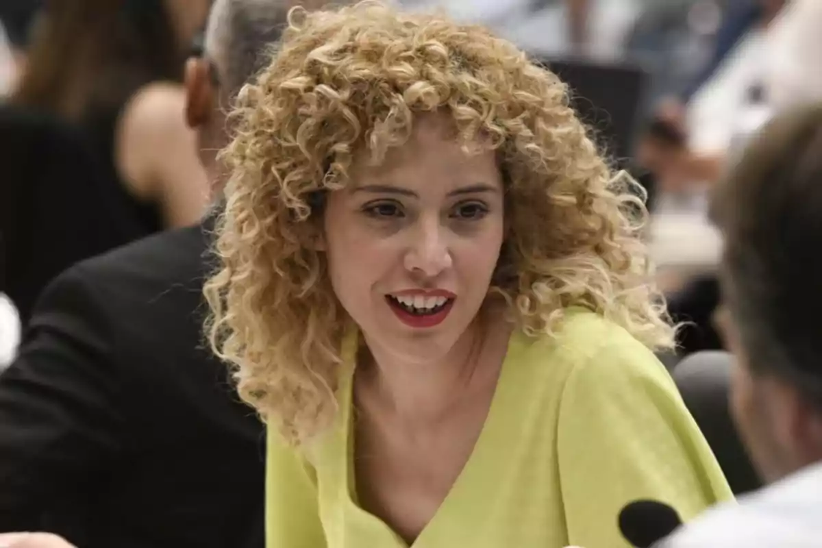 Blonde curly-haired woman wearing a yellow blouse talking in an office setting Blonde curly-haired woman wearing a yellow blouse talking in an office setting