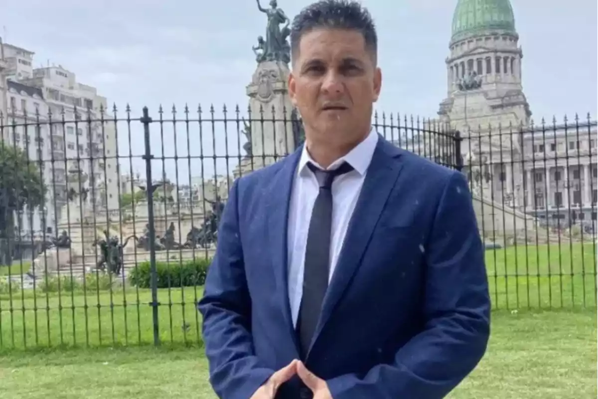 Short dark-haired man wearing a blue suit and black tie, posing in front of a fence with the Argentine National Congress in the background on a cloudy day. Short dark-haired man wearing a blue suit and black tie, posing in front of a fence with the Argentine National Congress in the background on a cloudy day.