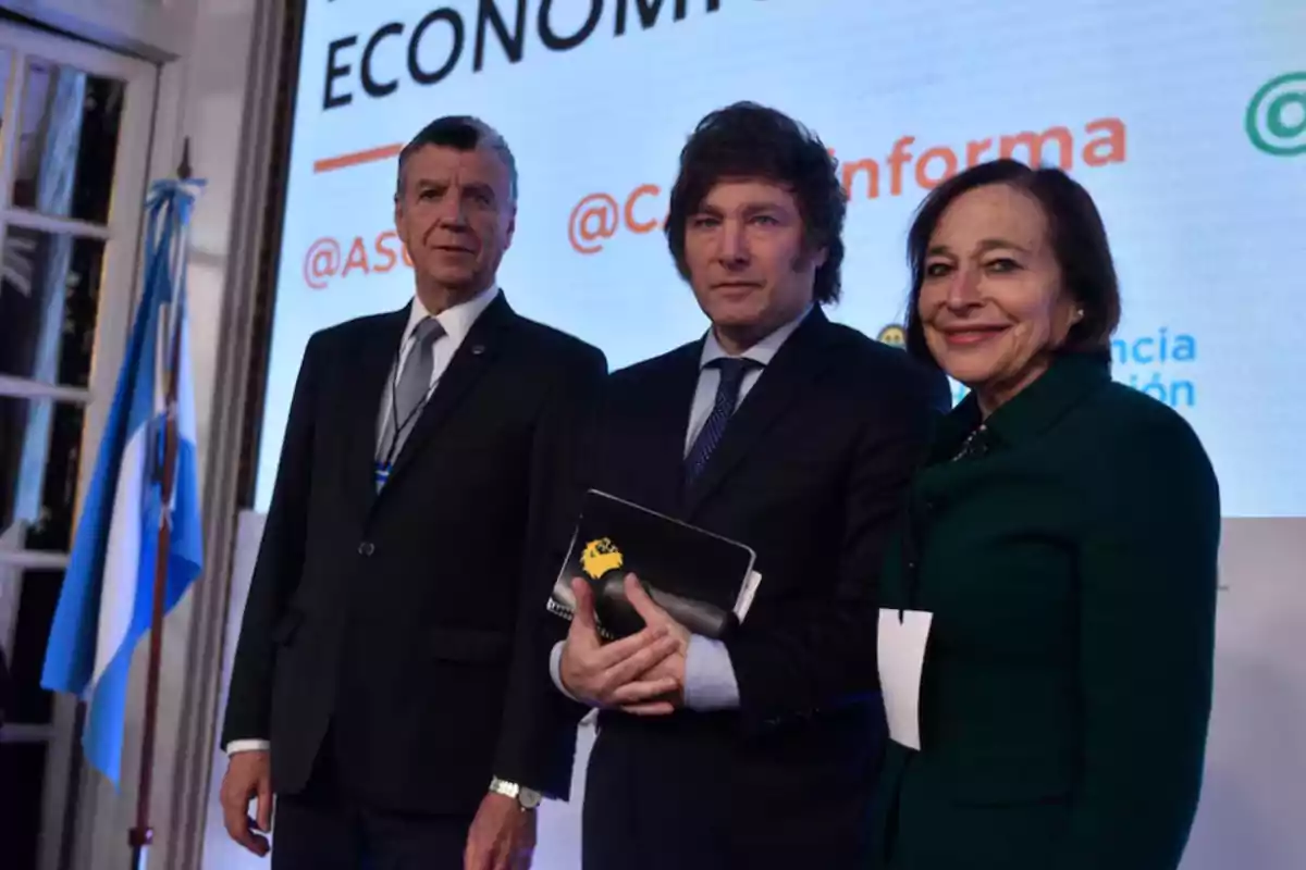 Three people dressed formally pose for a photo at an event with an Argentine flag and a screen with text in the background.