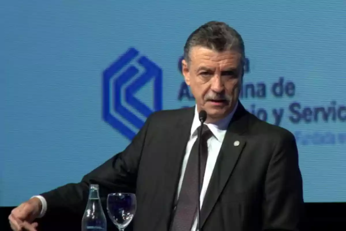 Man in a suit speaking at a podium with a microphone and a blue background with a logo and blurred text Man in a suit speaking at a podium with a microphone and a blue background with a logo and blurred text