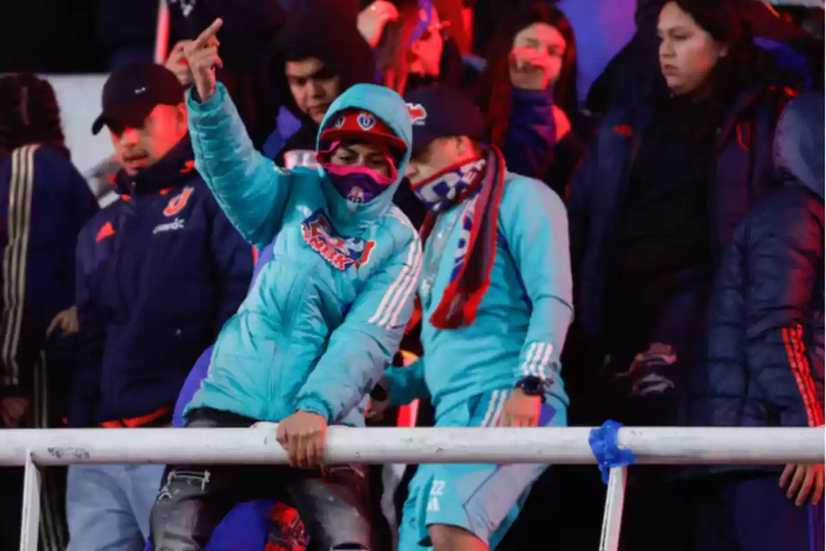 Aficionados de fútbol con ropa de invierno y bufandas en las gradas, una persona en primer plano hace un gesto obsceno con la mano Aficionados de fútbol con ropa de invierno y bufandas en las gradas, una persona en primer plano hace un gesto obsceno con la mano
