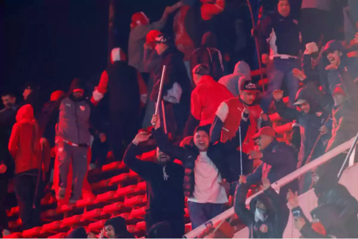 Aficionados en las gradas de un estadio con ropa roja y negra, algunos levantando los brazos y portando palos, en un ambiente de tensión y desorden Aficionados en las gradas de un estadio con ropa roja y negra, algunos levantando los brazos y portando palos, en un ambiente de tensión y desorden