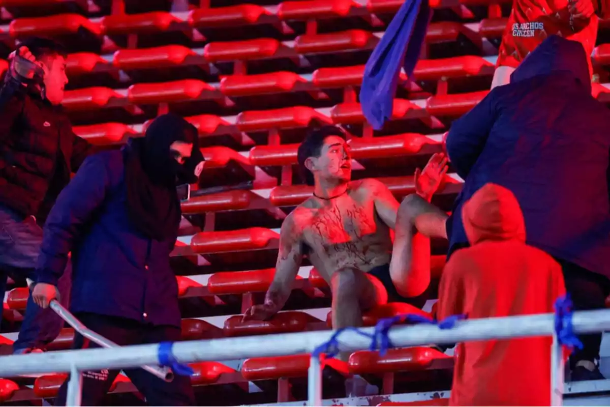 A young man, injured and covered in blood, is sitting in the stands of a stadium while several people surround him, some with their faces covered and carrying blunt objects, in a tense and violent atmosphere.