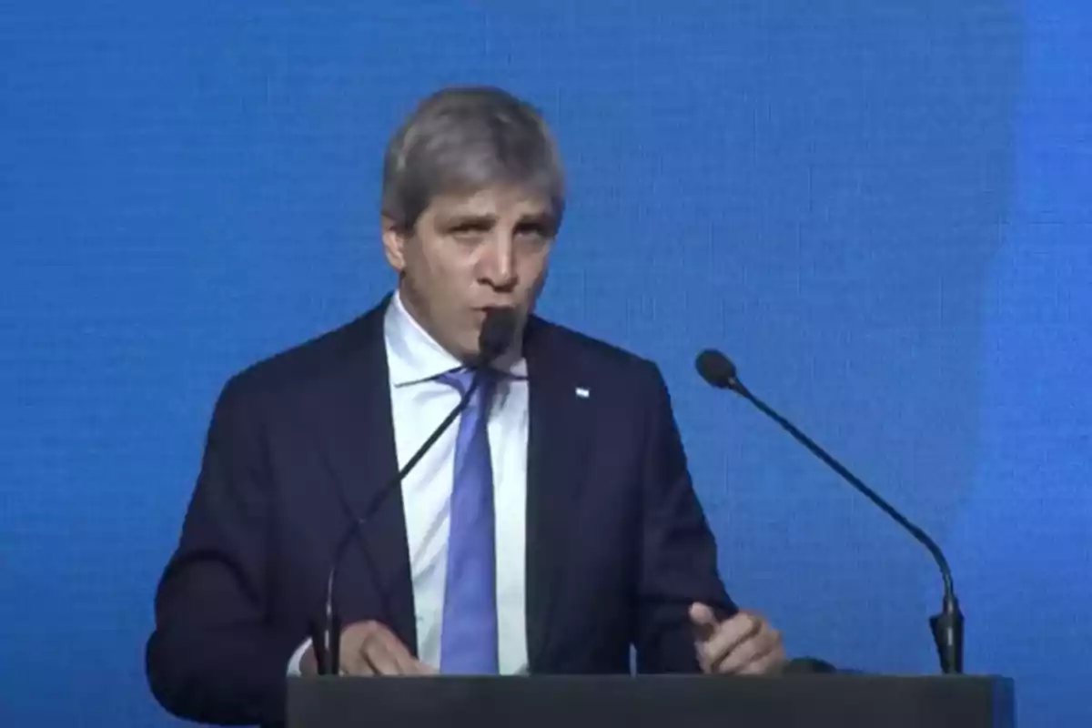 Gray-haired man in a dark suit and blue tie speaking at a podium in front of a blue background
