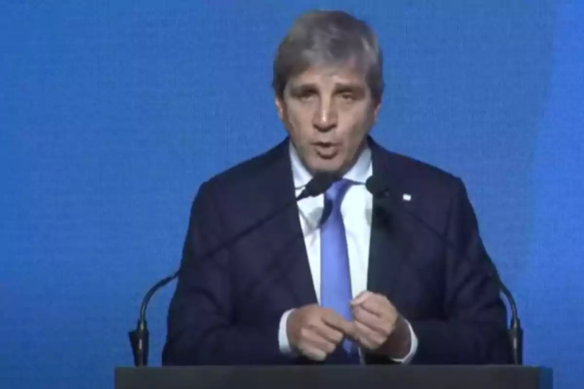 Gray-haired man in a dark suit and blue tie speaking in front of two microphones with a blue background Gray-haired man in a dark suit and blue tie speaking in front of two microphones with a blue background