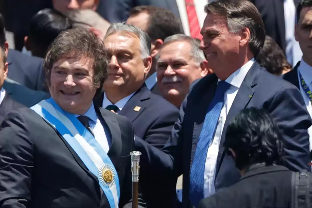 A man wearing a light blue and white presidential sash smiles, surrounded by other people in suits at an official outdoor event. A man wearing a light blue and white presidential sash smiles, surrounded by other people in suits at an official outdoor event.