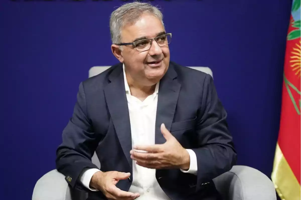 Gray-haired man with glasses sitting on a chair with a blue background and a flag beside him while gesturing with his hand Gray-haired man with glasses sitting on a chair with a blue background and a flag beside him while gesturing with his hand