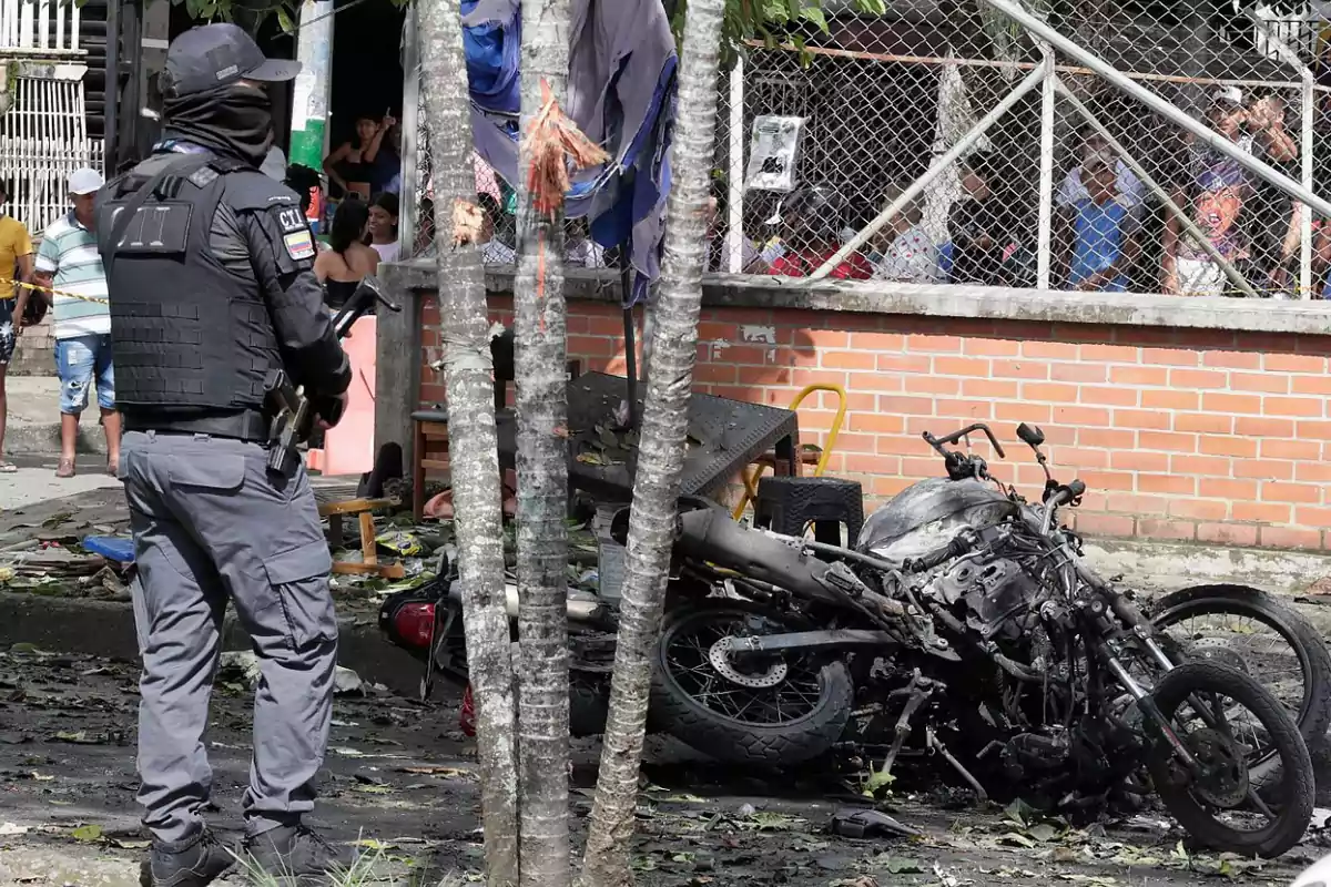 Un agente de policía observa una motocicleta calcinada en una calle mientras varias personas miran la escena desde detrás de una reja Un agente de policía observa una motocicleta calcinada en una calle mientras varias personas miran la escena desde detrás de una reja