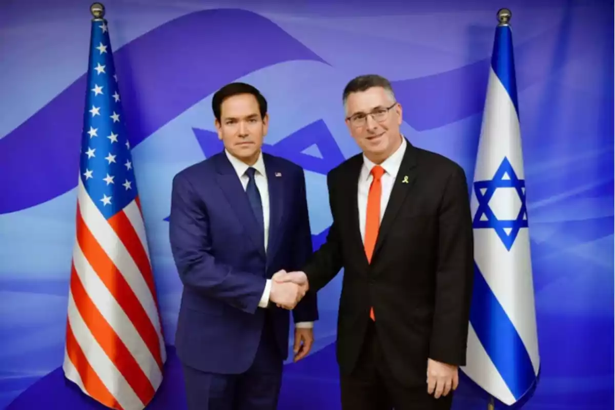 Two men in suits shake hands in front of the flags of the United States and Israel.