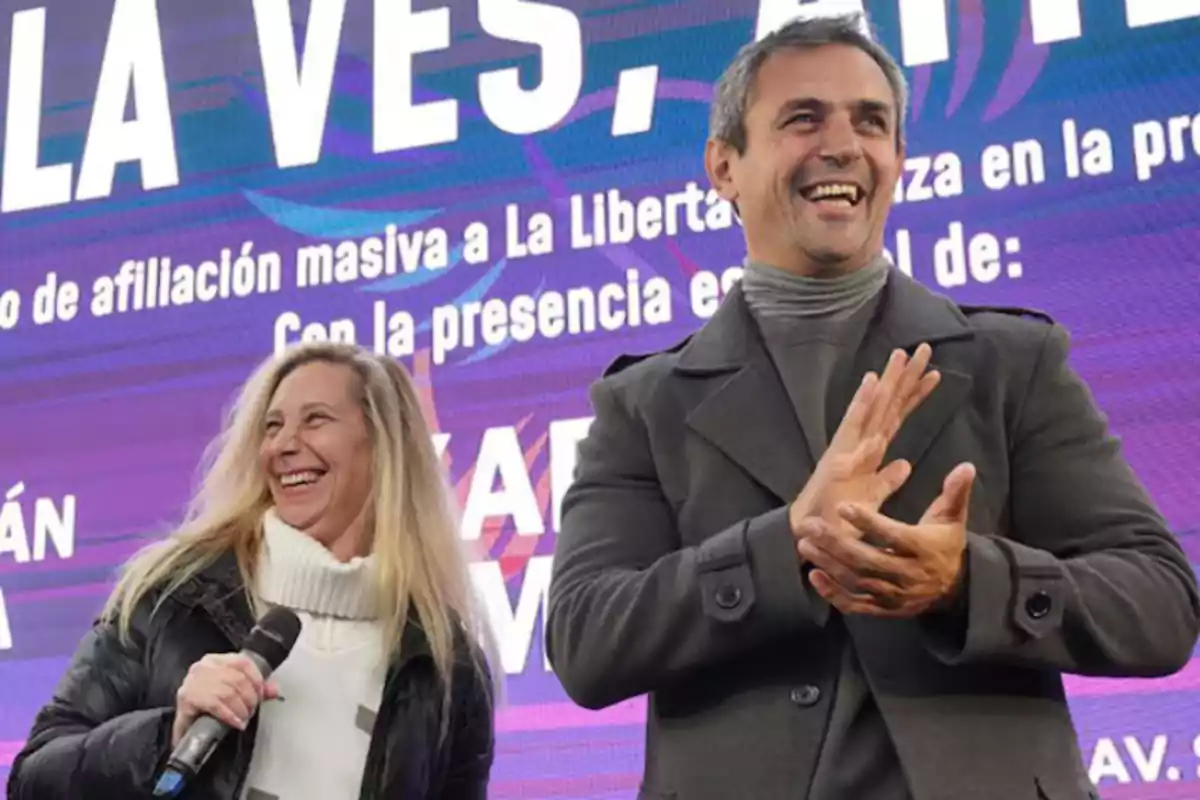 Two people are smiling on a stage in front of a screen with large, colorful letters; one of them is holding a microphone and the other is clapping.