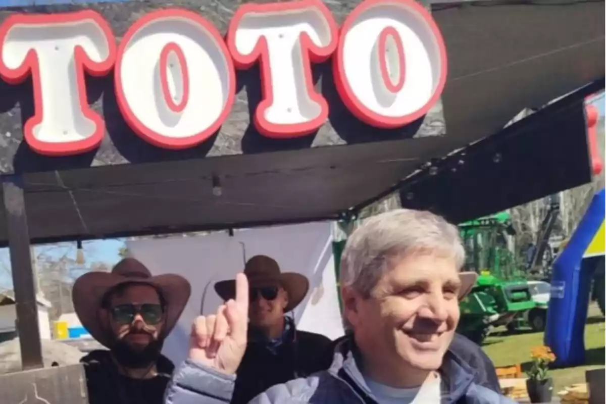 A smiling man points his finger at a large sign that says TOTO while two people wearing hats stand behind him. A smiling man points his finger at a large sign that says TOTO while two people wearing hats stand behind him.