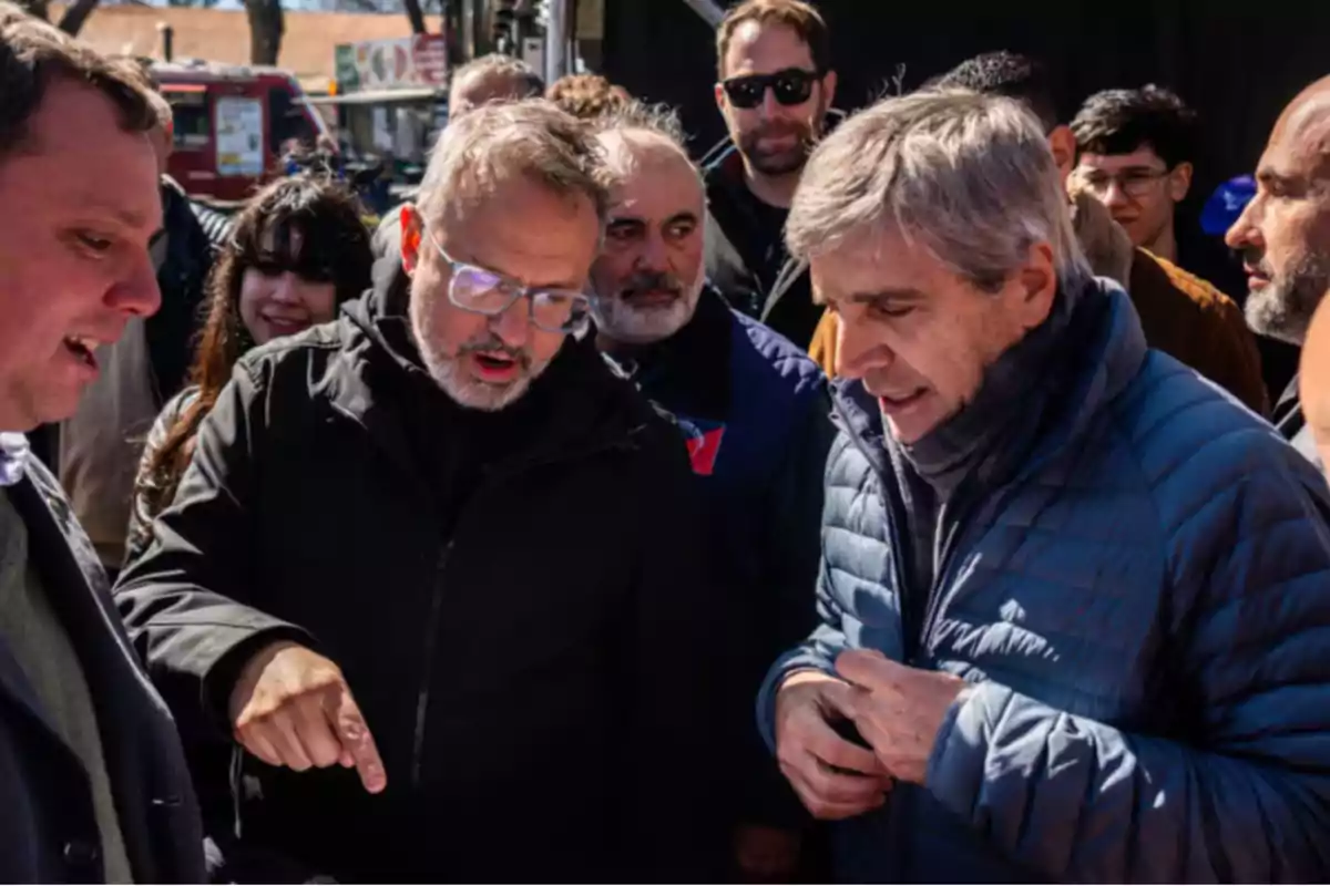 A group of people is talking outdoors on a sunny day; some of them are wearing warm clothes and seem focused on a discussion.