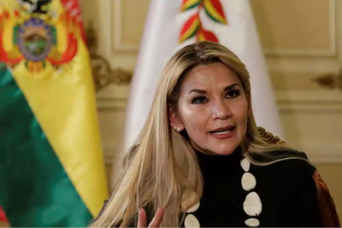 Mujer de cabello rubio hablando frente a dos banderas en un entorno formal Mujer de cabello rubio hablando frente a dos banderas en un entorno formal