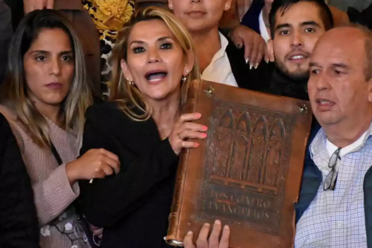 Mujer rubia en el centro de un grupo de personas sosteniendo un gran libro antiguo de tapa marrón con relieves y la inscripción Los Cuatro Evangelios