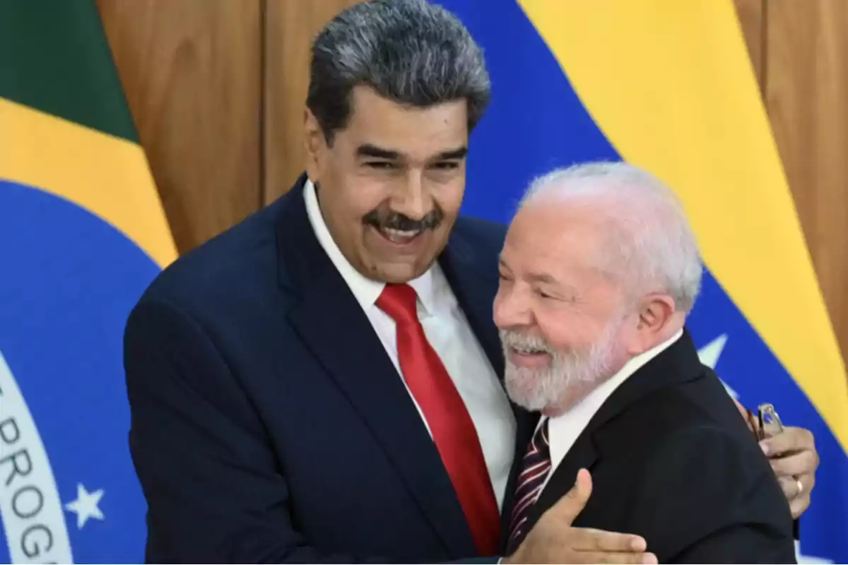Two men in suits embrace, smiling, in front of the flags of Brazil and Venezuela.