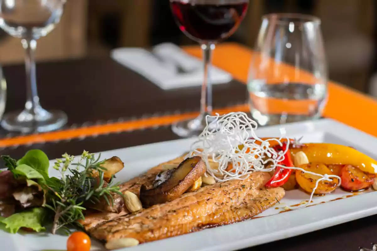 Plato gourmet de salmón a la plancha acompañado de vegetales asados, ensalada fresca, decoración de fideos crujientes y una copa de vino tinto al fondo
